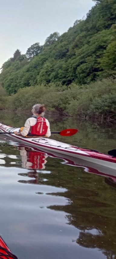 Das rotweiße Kajak von der Seite sichtbar auf der Lahn und darin eine Frau mit Schwimmweste.